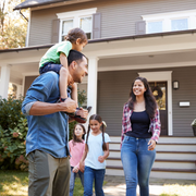 Family standing in front of a house, with a man carrying a child on his shoulders and two children nearby.