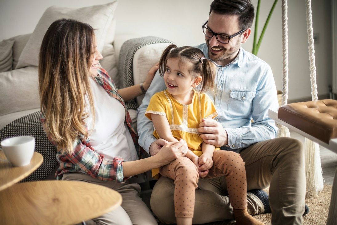 Family sitting together on a couch, smiling and holding a small child in a bright living room.