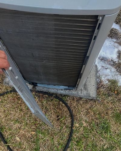 Close-up of an HVAC unit outdoors, with a hand holding a metal tool beside the condenser coil.