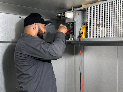 Technician wiring equipment inside a metal cabinet with a yellow meter and red-black leads.