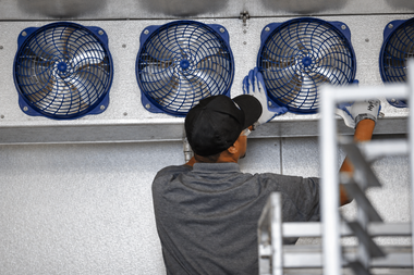 Worker in a black cap adjusts blue wall-mounted fans in an industrial room.