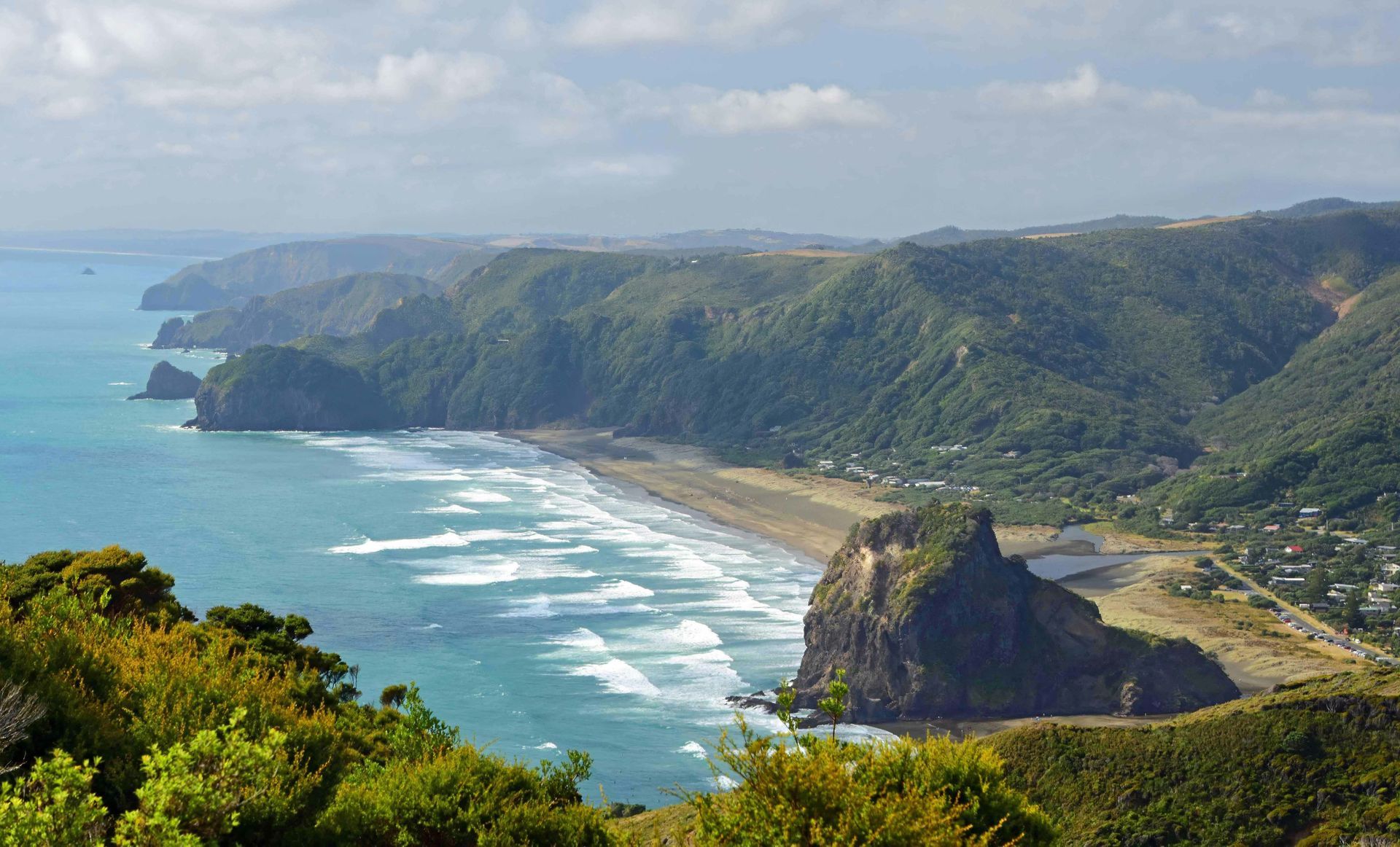 View of a rugged coastline with dark sand beach, green hills, and turquoise ocean under a cloudy sky.