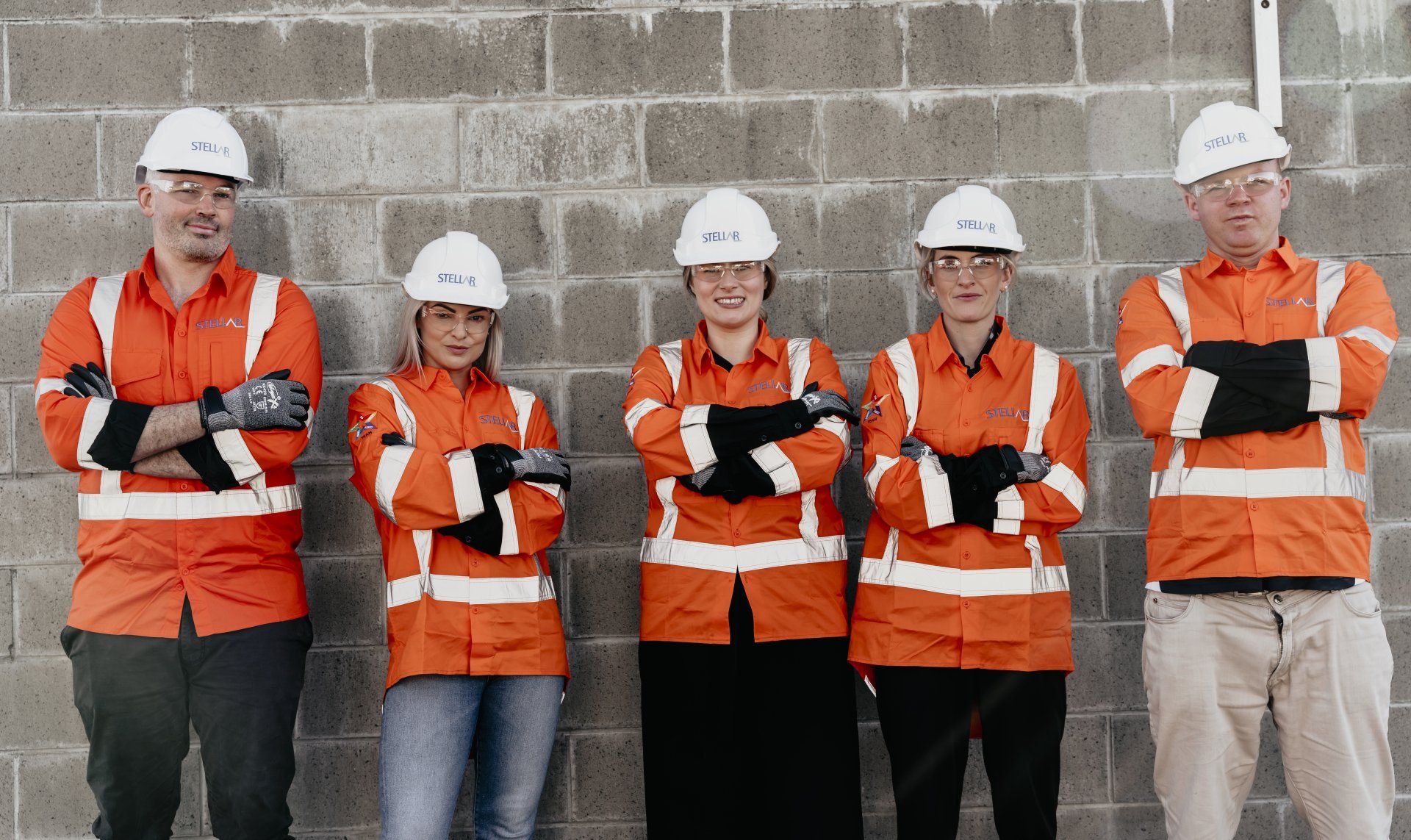 Five construction workers in orange vests and hard hats stand in front of a gray brick wall, arms crossed.