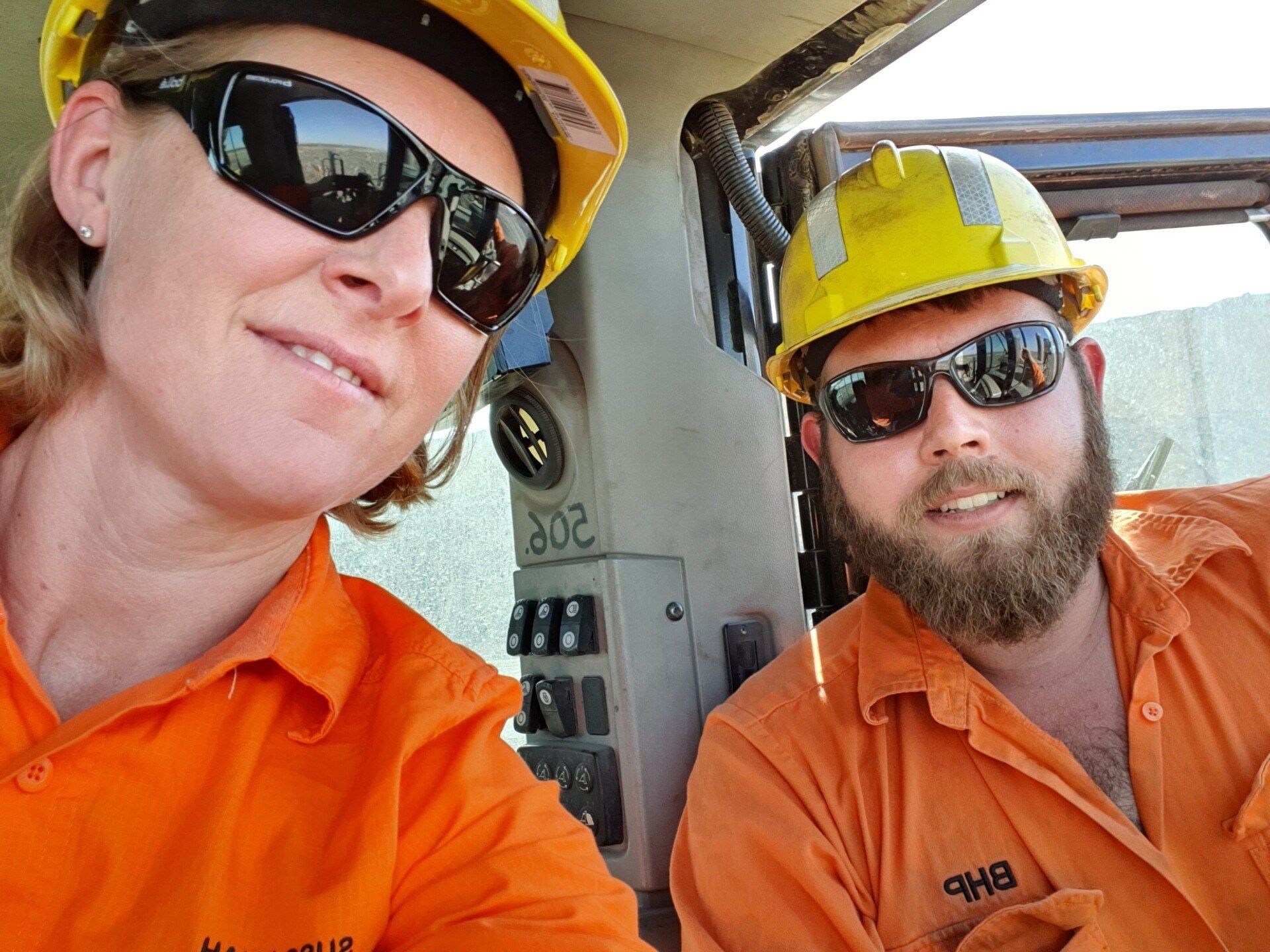 Two people in orange work uniforms and hard hats, wearing sunglasses, smiling for a selfie in a vehicle.