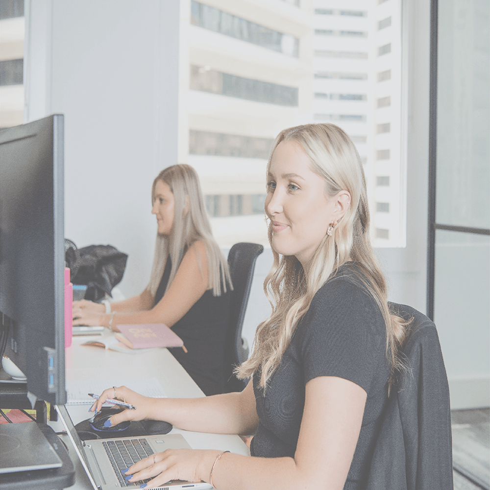 Two blond women work at desks in a bright office, typing and looking at computer screens.
