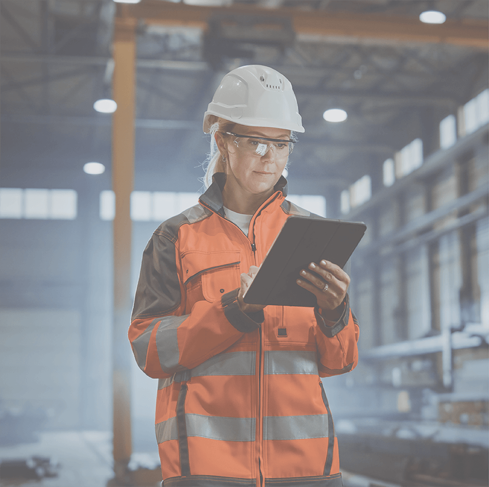 Woman in hard hat and safety vest using tablet in industrial setting.
