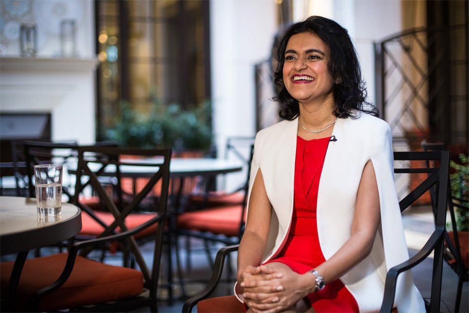 Woman in red dress and white cape blazer smiling, seated in outdoor cafe.