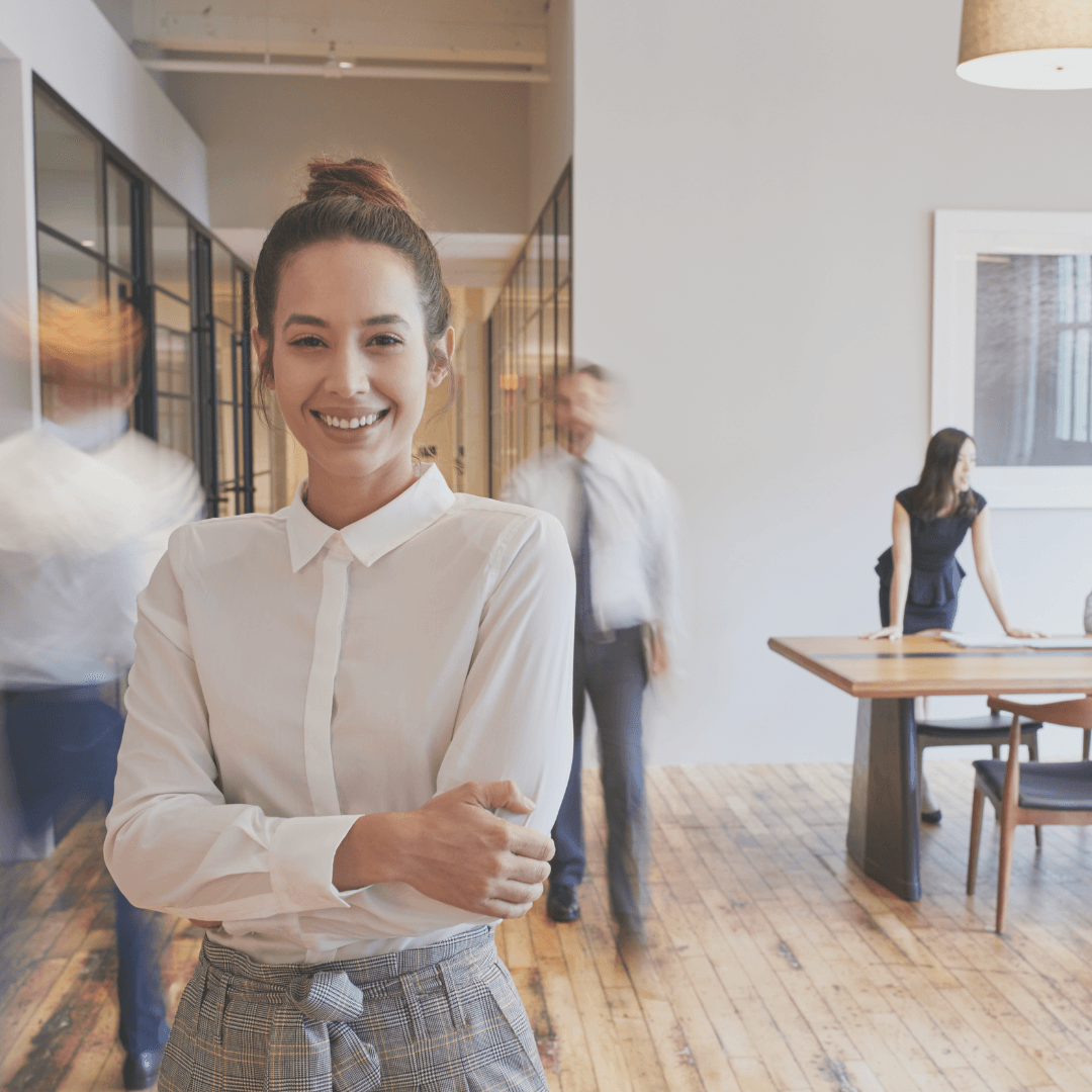 Woman in office, arms crossed, smiling at camera; blurred coworkers.