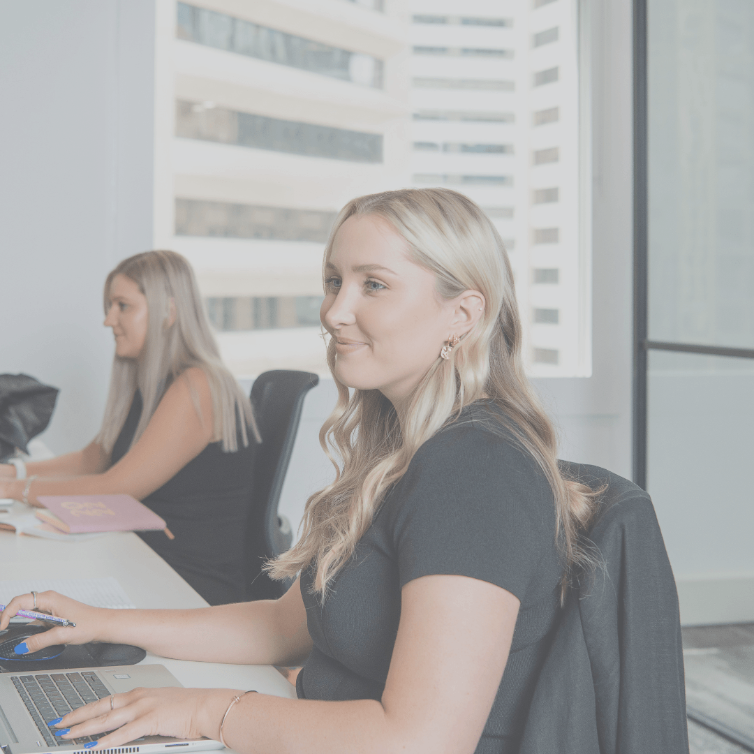 Two blonde women work at desks in a modern office, one smiling at a computer.