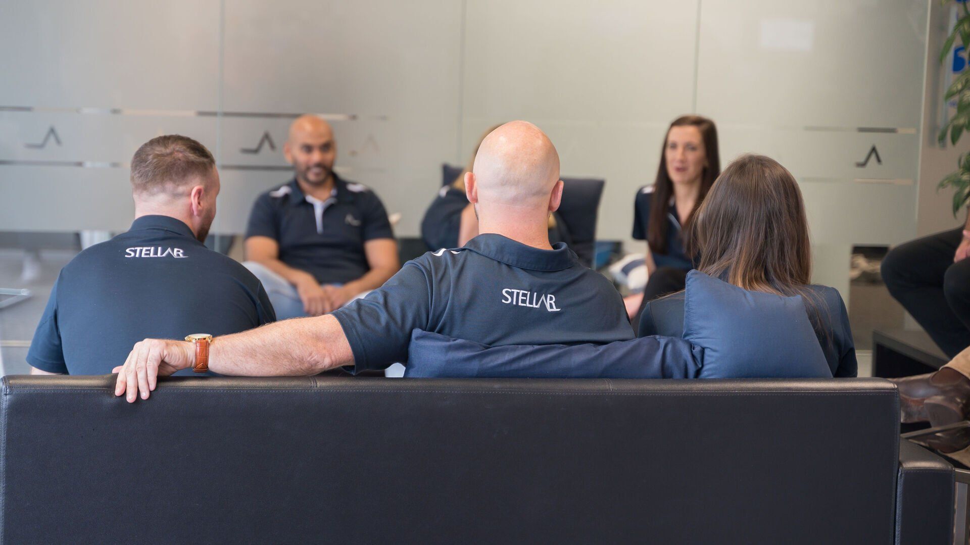 People in a modern office setting, sitting on a black couch, having a meeting.