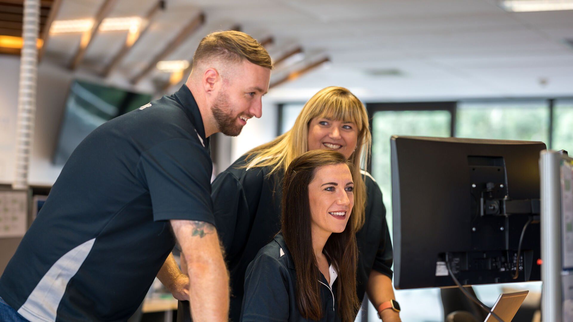 Three coworkers looking at a computer screen, smiling. Office setting, dressed in matching shirts.
