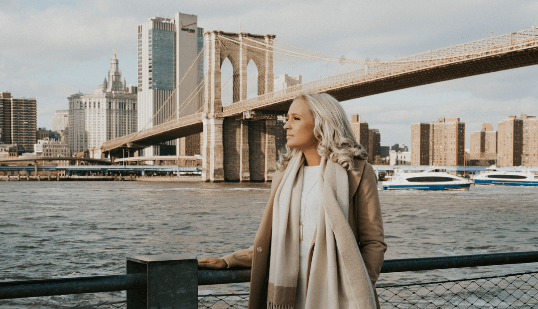 Woman with white hair looks towards NYC skyline and Brooklyn Bridge.