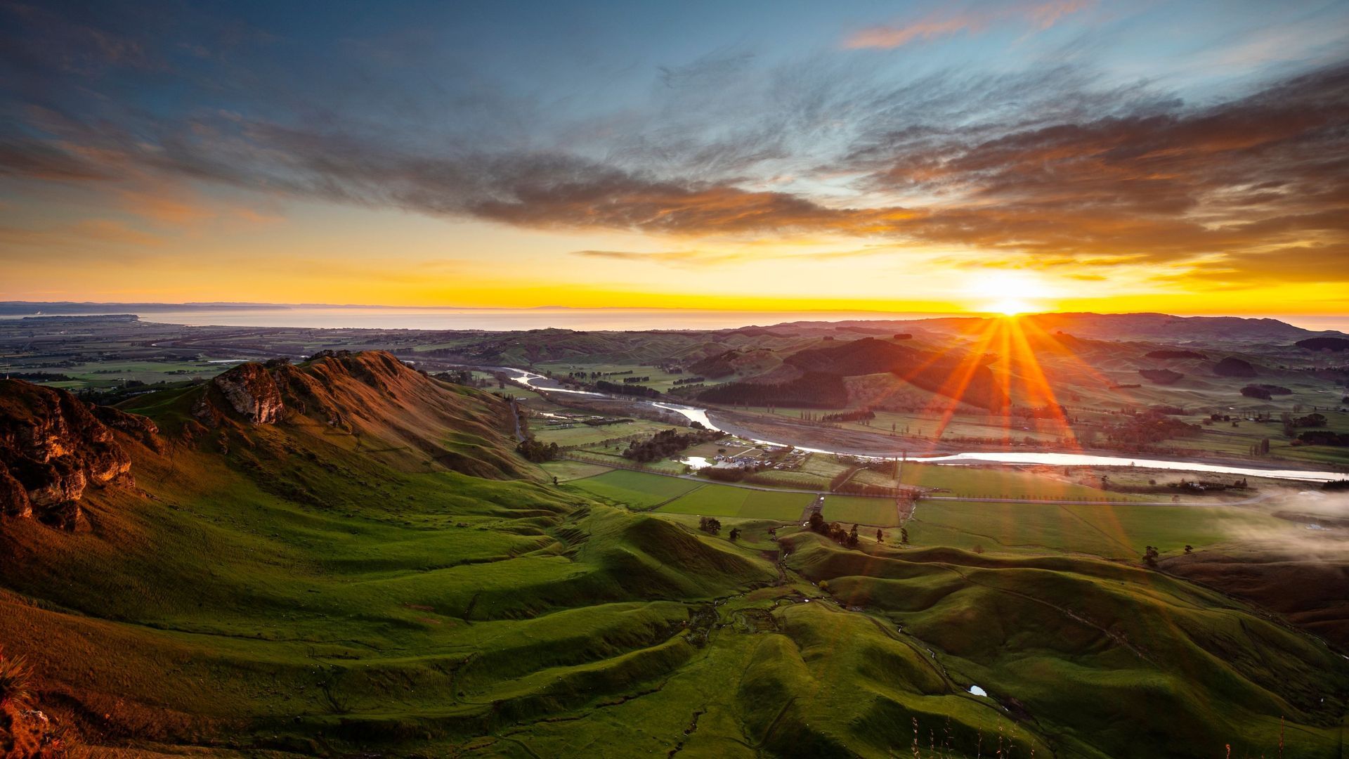 Rolling green hills and a river at sunrise. Bright sunburst illuminates a vibrant landscape.