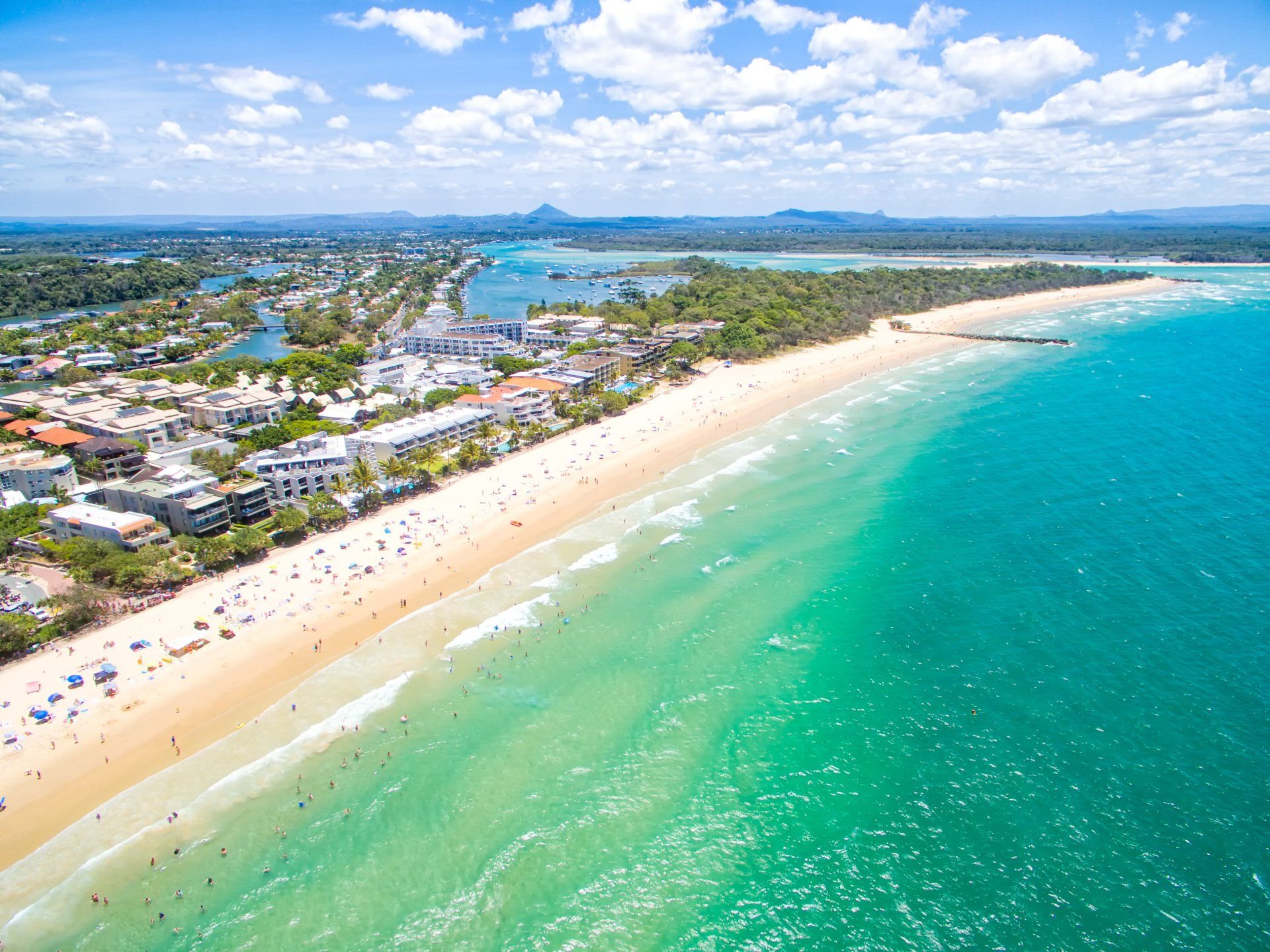 Aerial view of a beach with turquoise water, golden sand, buildings, trees, and people on a sunny day.