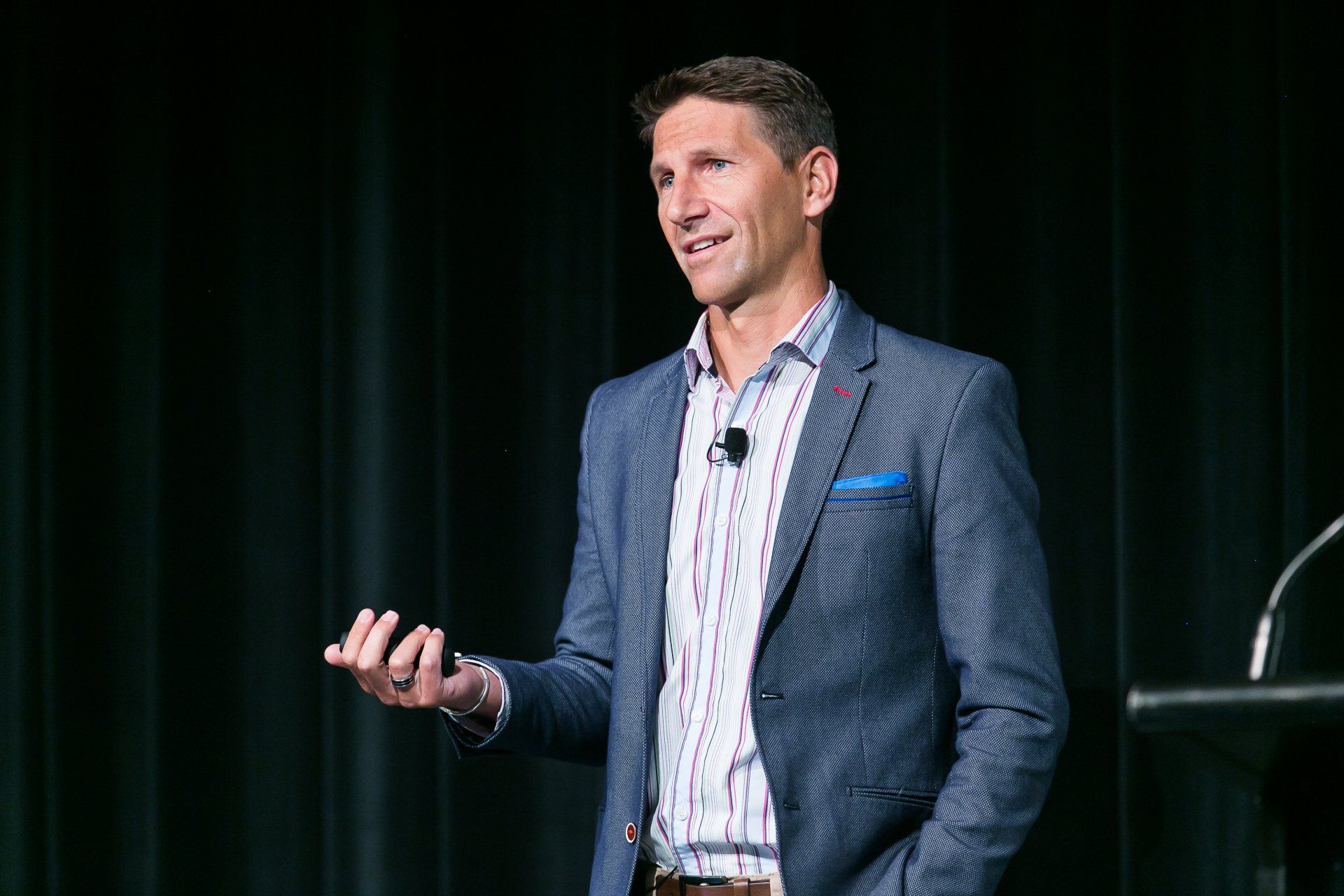 Man in a blue blazer speaks at a conference, holding a small object, standing in front of a dark curtain.