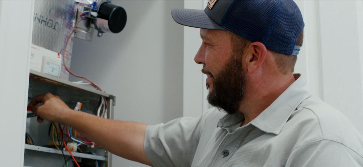 A man with a beard and hat works on electrical wiring inside a metal box in a room.