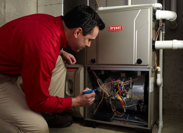 Man in red shirt inspecting a furnace, using a tool, indoors.
