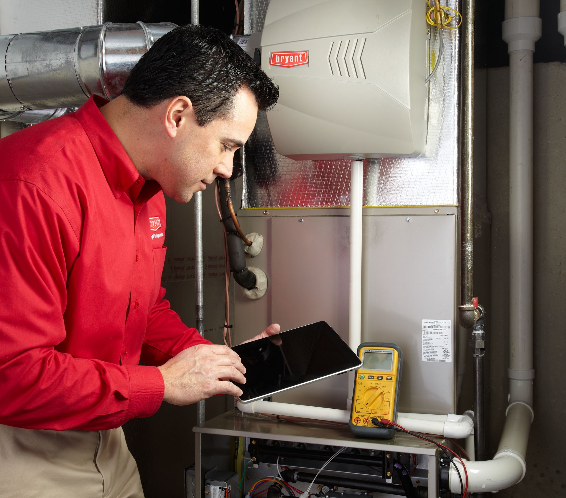 HVAC technician in red shirt examines a furnace, using a tablet and multimeter in a utility room.
