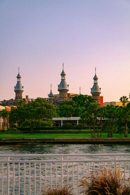 Riverfront view of a white-domed mosque at sunset, with trees and a bridge railing in the foreground
