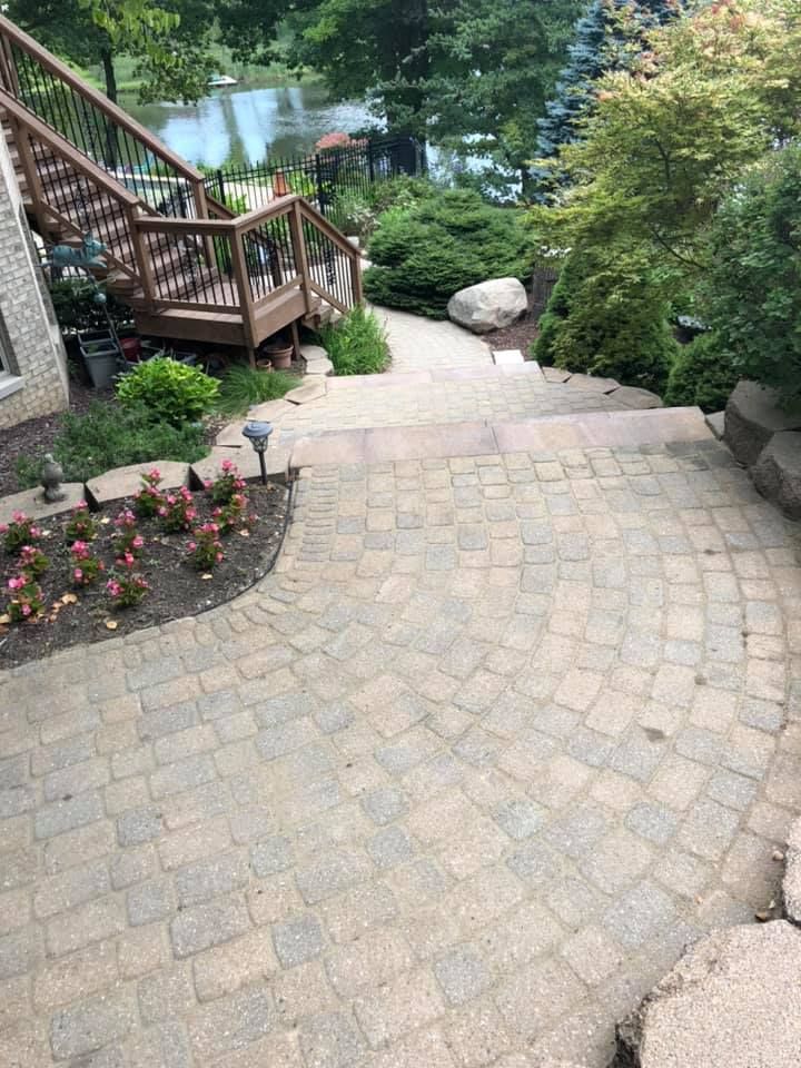 Paver stone patio path beside wooden stairs, garden flowers, trees, and a view of water beyond