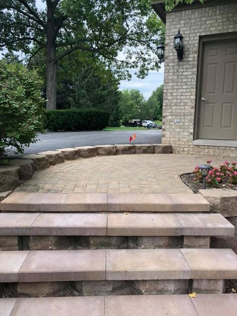 Stone steps leading to a brick patio beside a house, with trees and a street in the background.