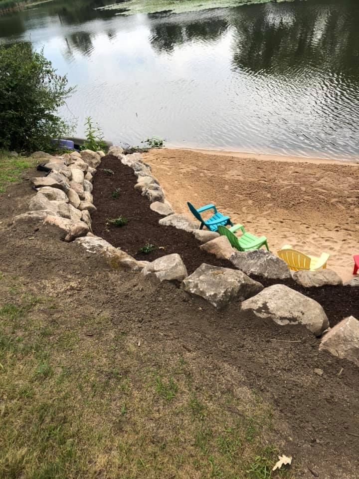 Shoreline garden bed with rocks, soil, and small plants beside a sandy lake edge with colorful chairs.