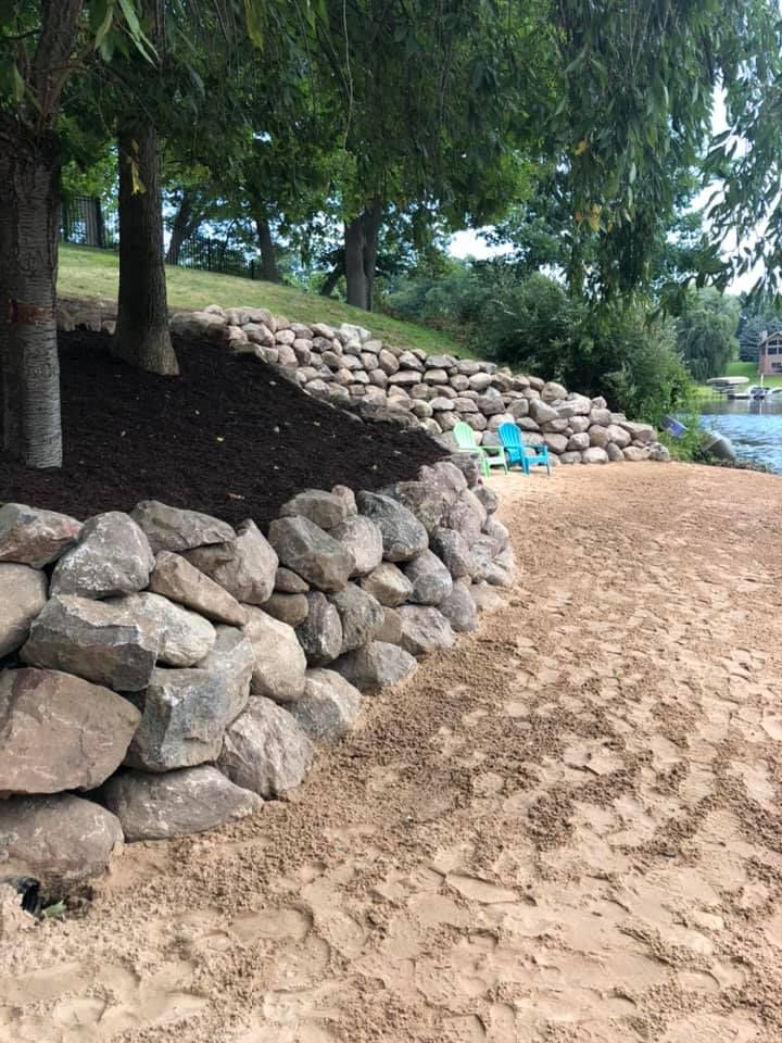 Stone retaining wall beside a sandy path under trees, with a glimpse of water in the background