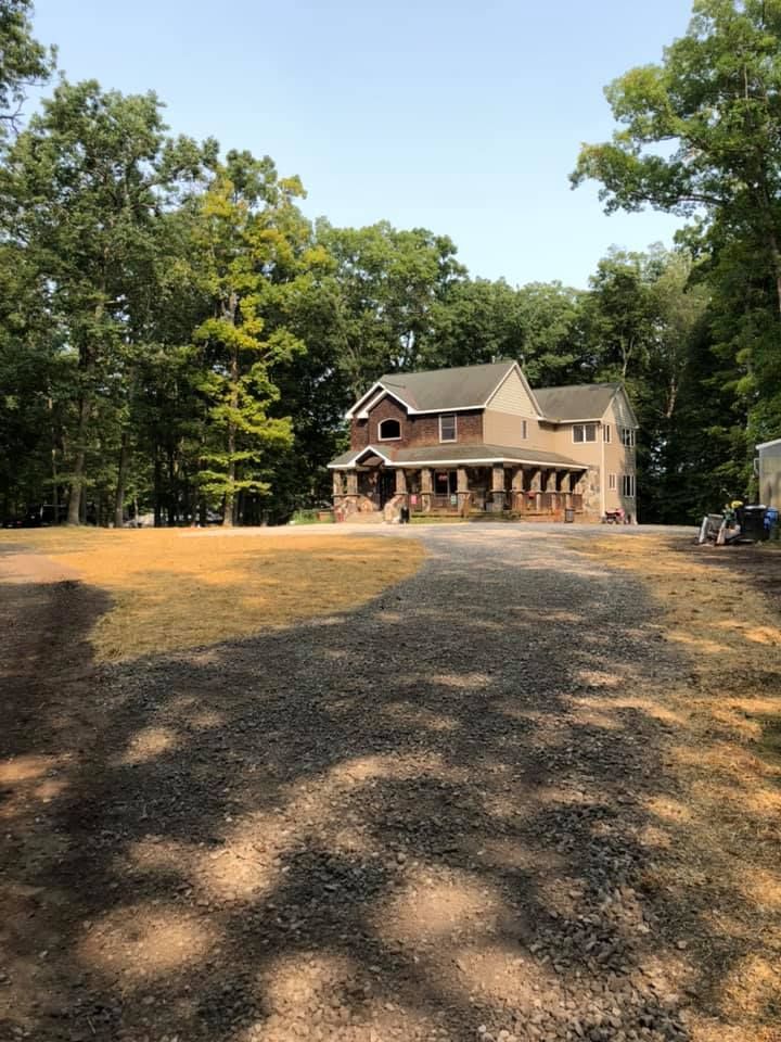 Brick house with porch and green shutters in a wooded yard, viewed from a gravel driveway