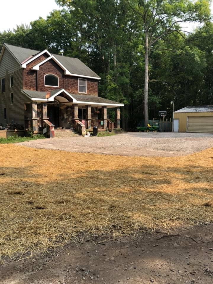 Brick house with covered porch and large gravel driveway beside a detached garage, surrounded by trees