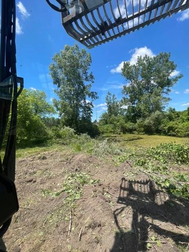 Cleared dirt lot with trees under a bright blue sky, viewed from heavy machinery arm in the foreground