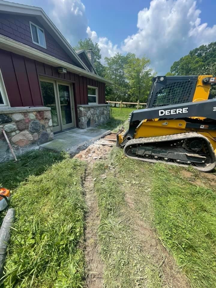 John Deere skid steer beside house, grading dirt along a stone-and-red siding home under a partly cloudy sky