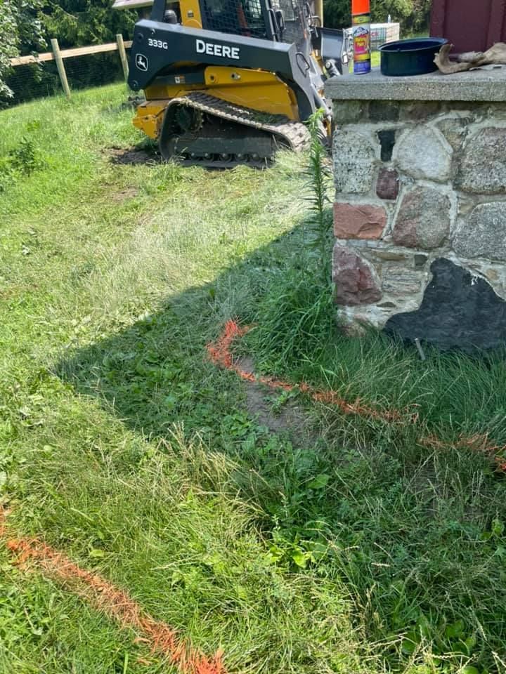 Yellow John Deere skid steer mows grass beside a stone wall in a sunny yard.