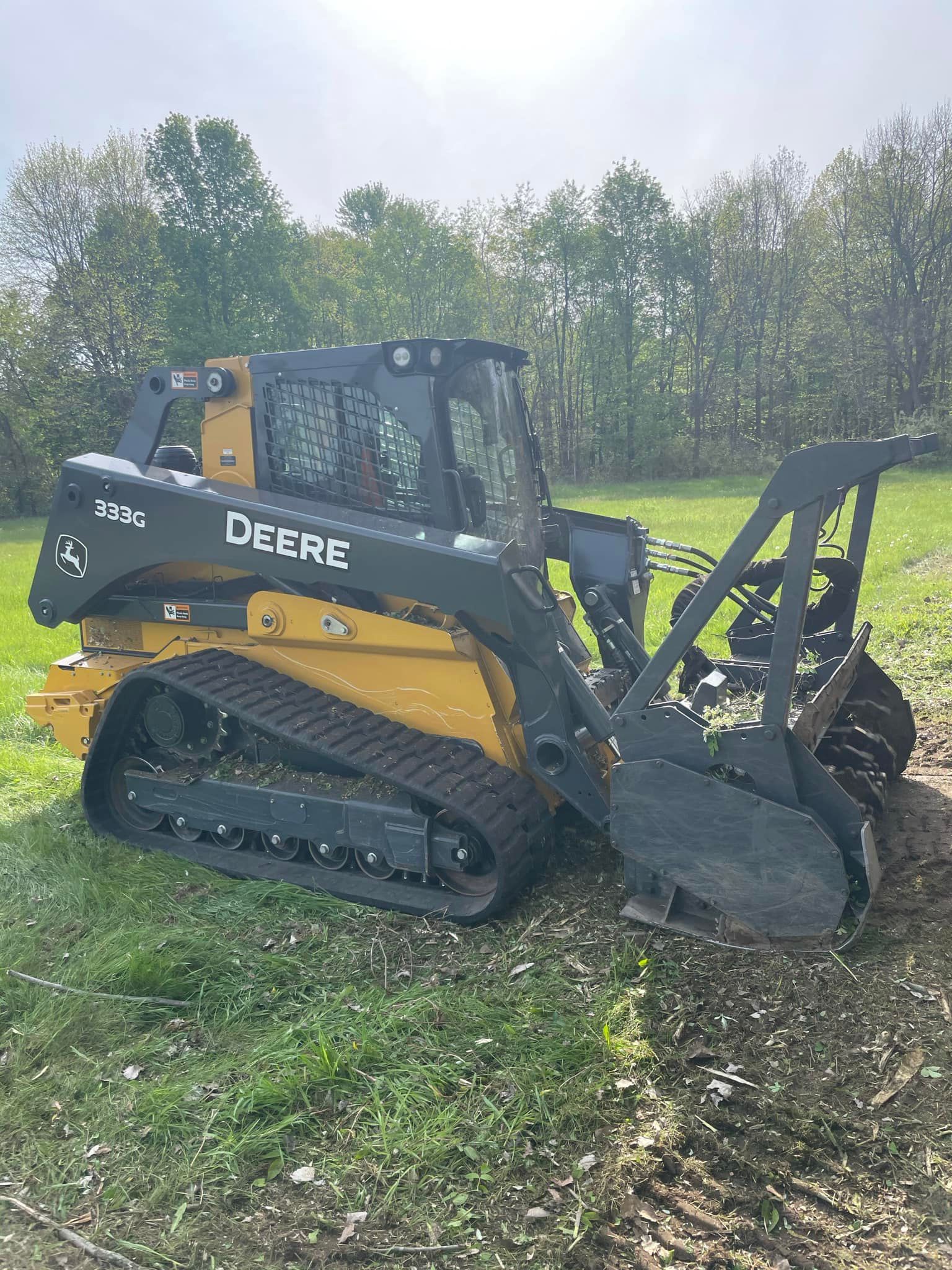 Yellow John Deere compact track loader with front attachment in a grassy field near trees.