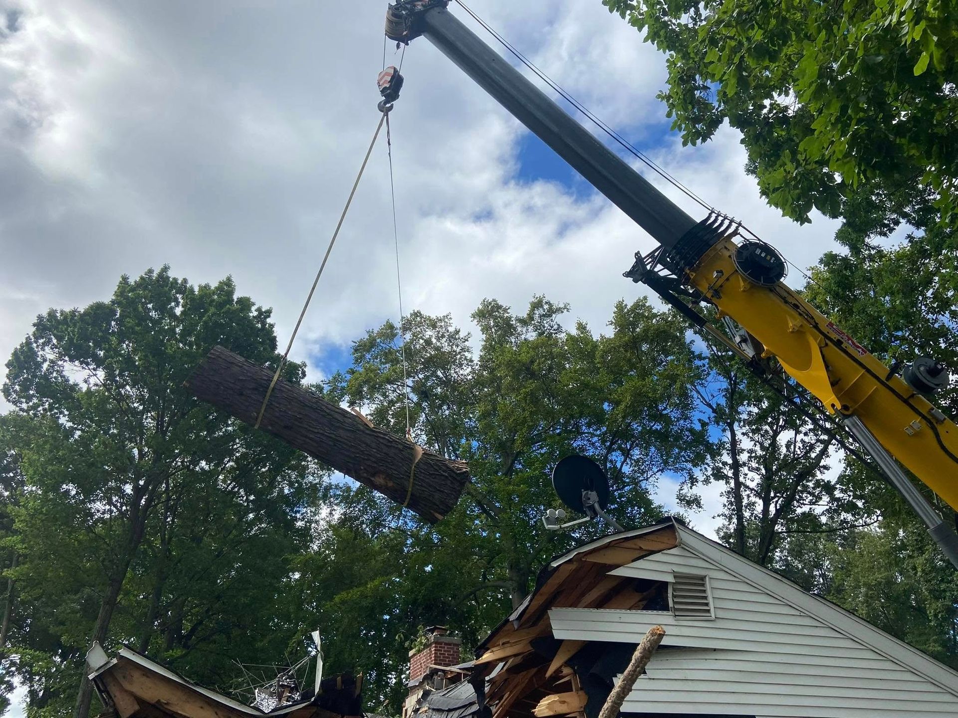 Crane lifting a large fallen tree limb over a damaged roof in a residential yard
