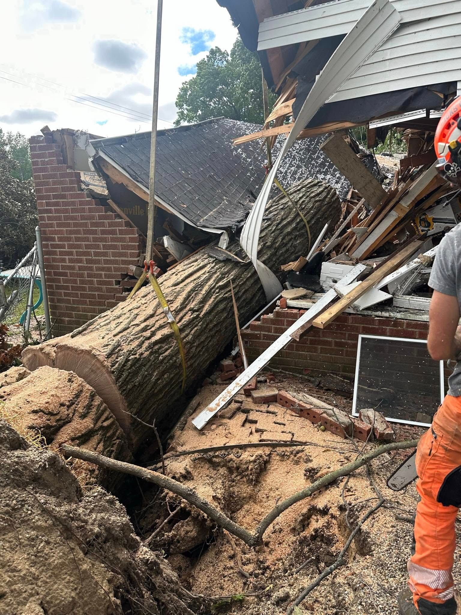 Fallen tree crushed a house roof and debris; a worker in orange stands at the right.