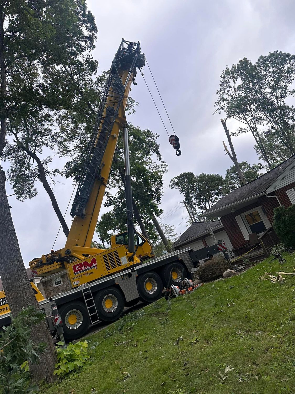 Yellow crane lifting debris near a house in a wooded yard, with a worker suspended on the hook.