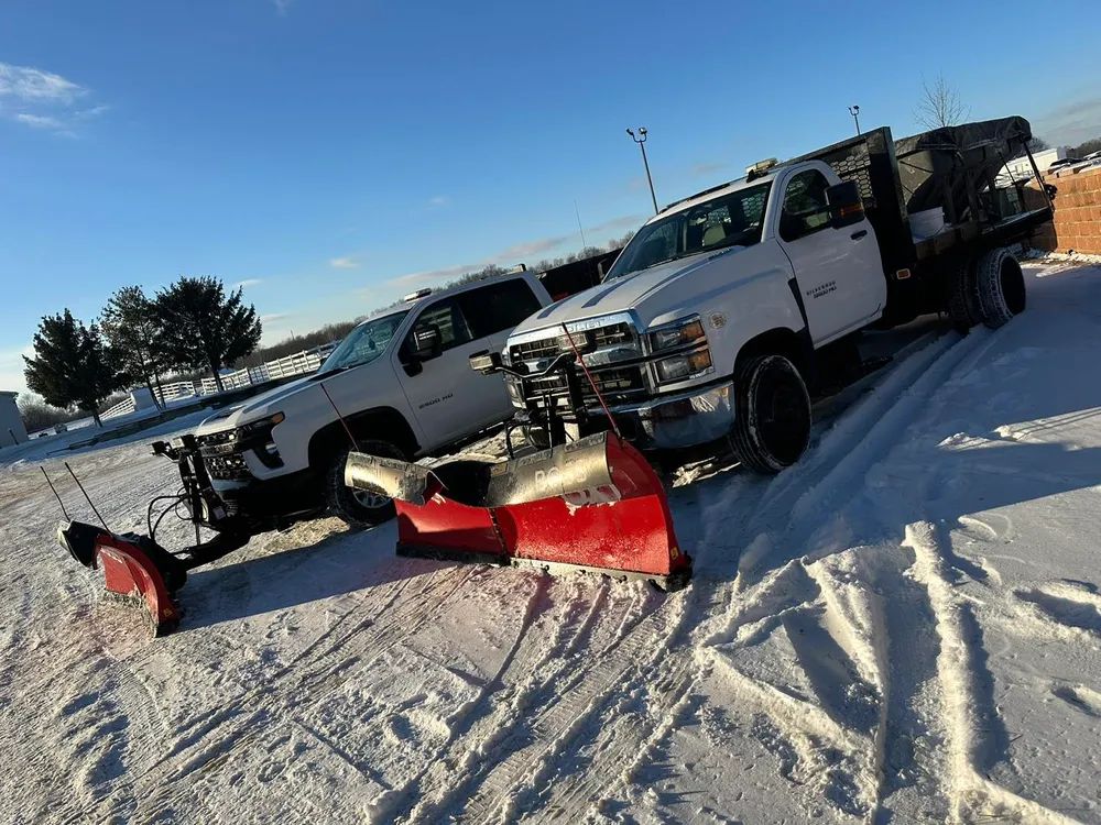 Two snowplow trucks with red blades parked on a snowy lot under a clear blue sky