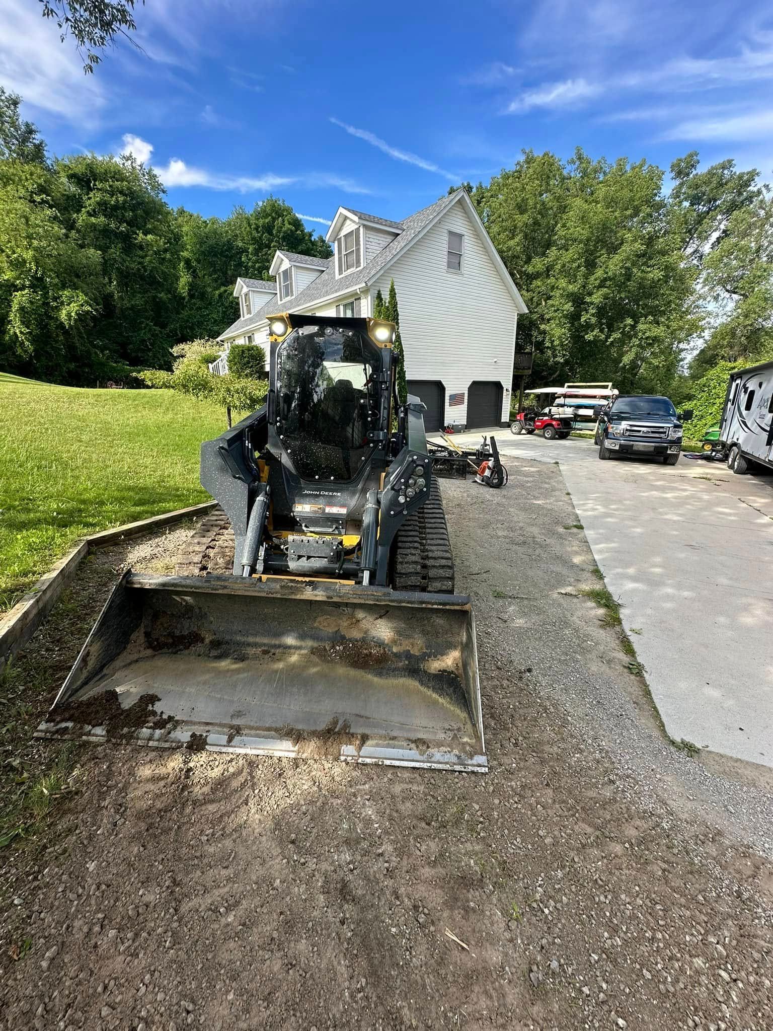 Bulldozer with raised blade on a driveway beside a white house and parked cars, under a sunny blue sky