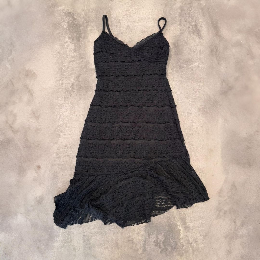 Vestido de encaje negro con tirantes finos, volantes en capas y dobladillo acampanado sobre un fondo neutro.