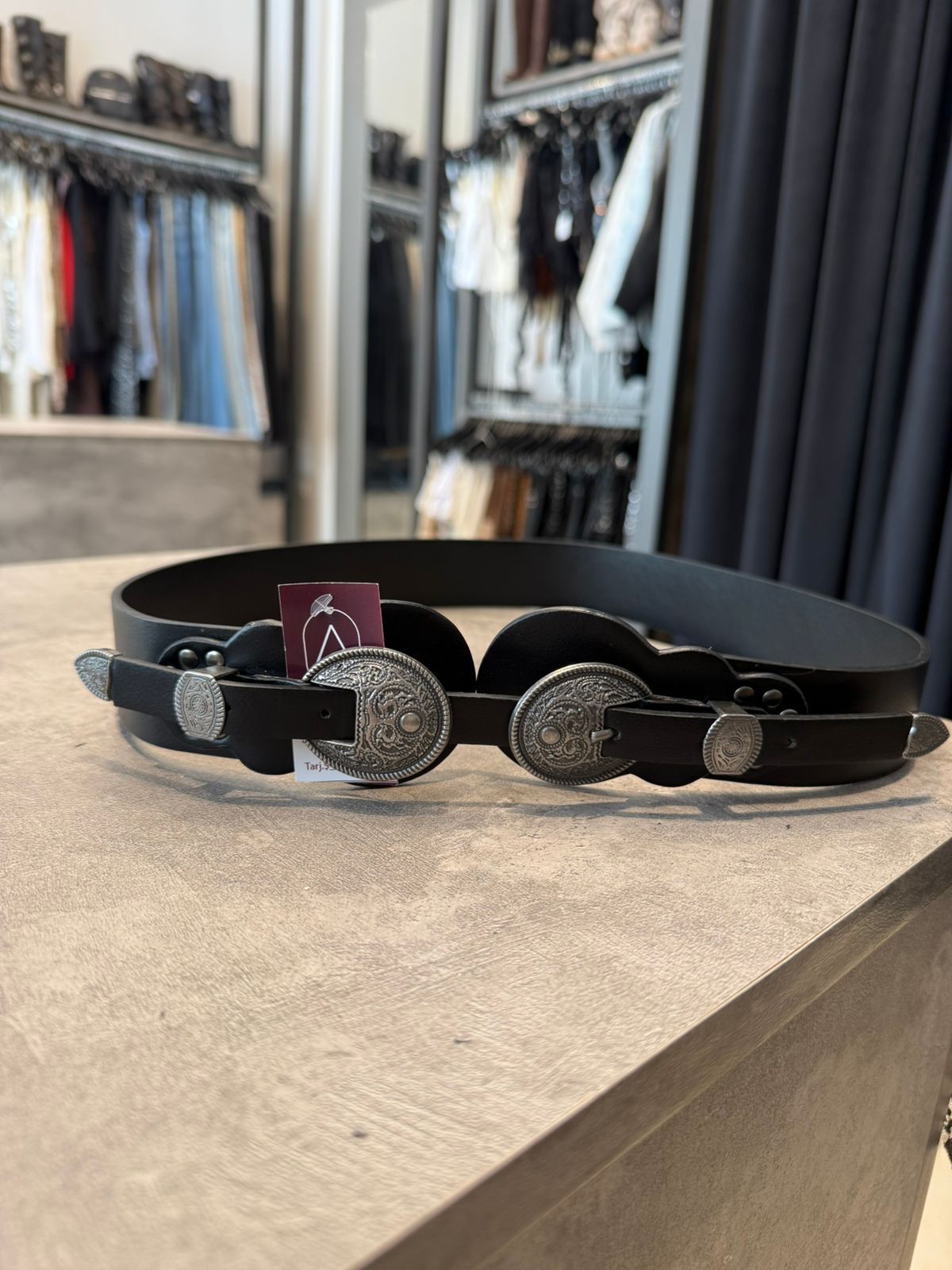 Cinturón de cuero negro con hebillas decorativas plateadas en expositor de tienda.