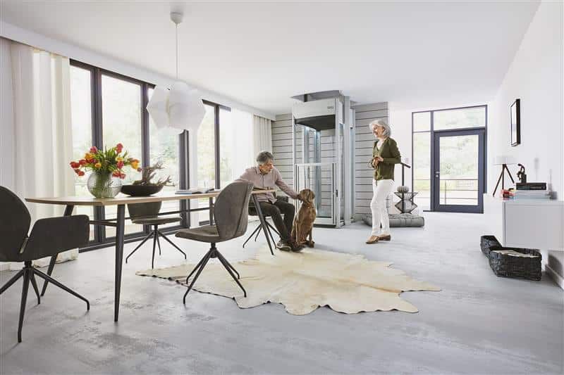 Modern open living room with large windows, dining table, chairs, rug, and two people standing and seated. Modern open living room with large windows, dining table, chairs, rug, and two people standing and seated.