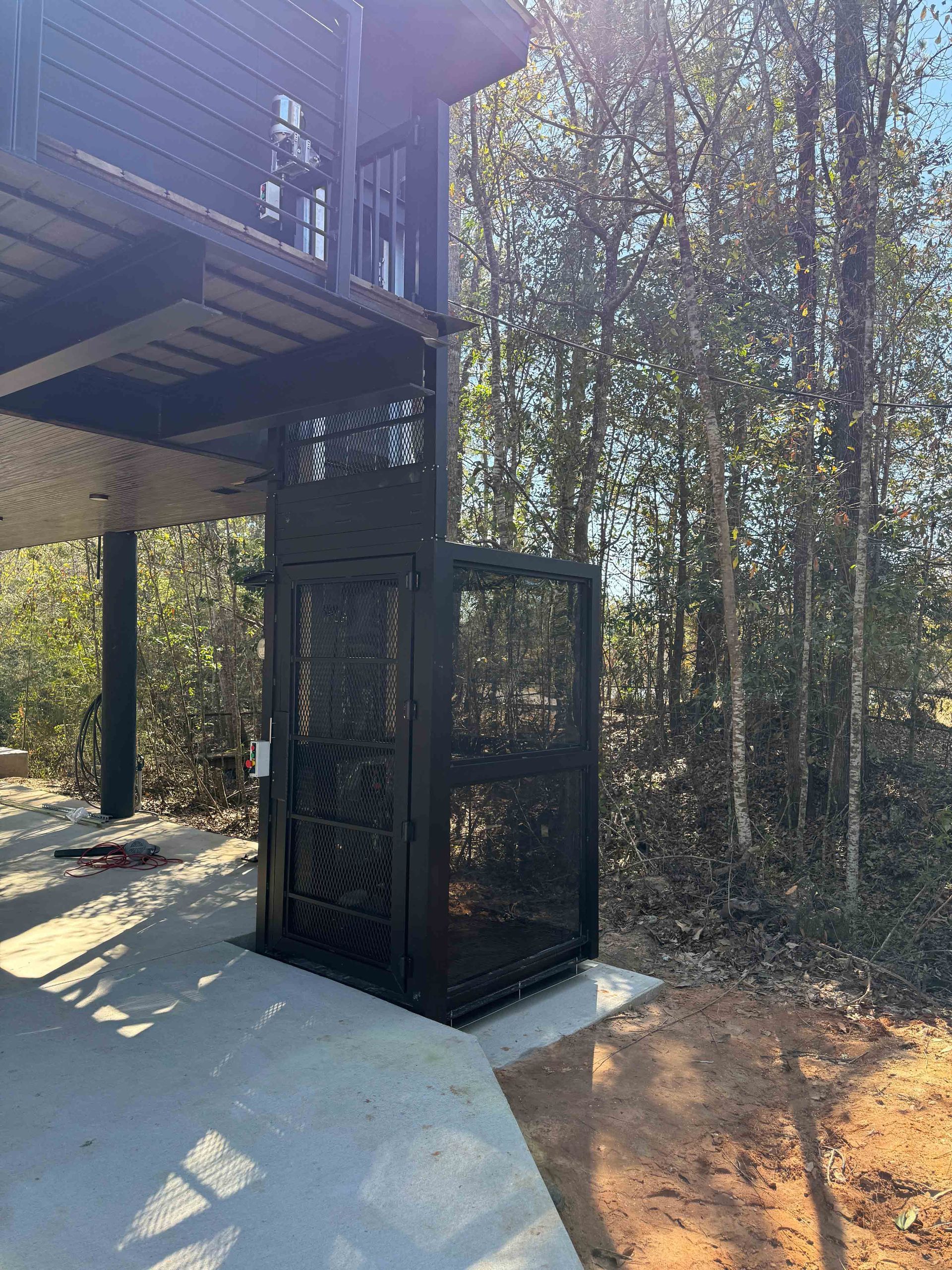 Black metal outdoor elevator beside a raised deck, near a concrete path and wooded trees