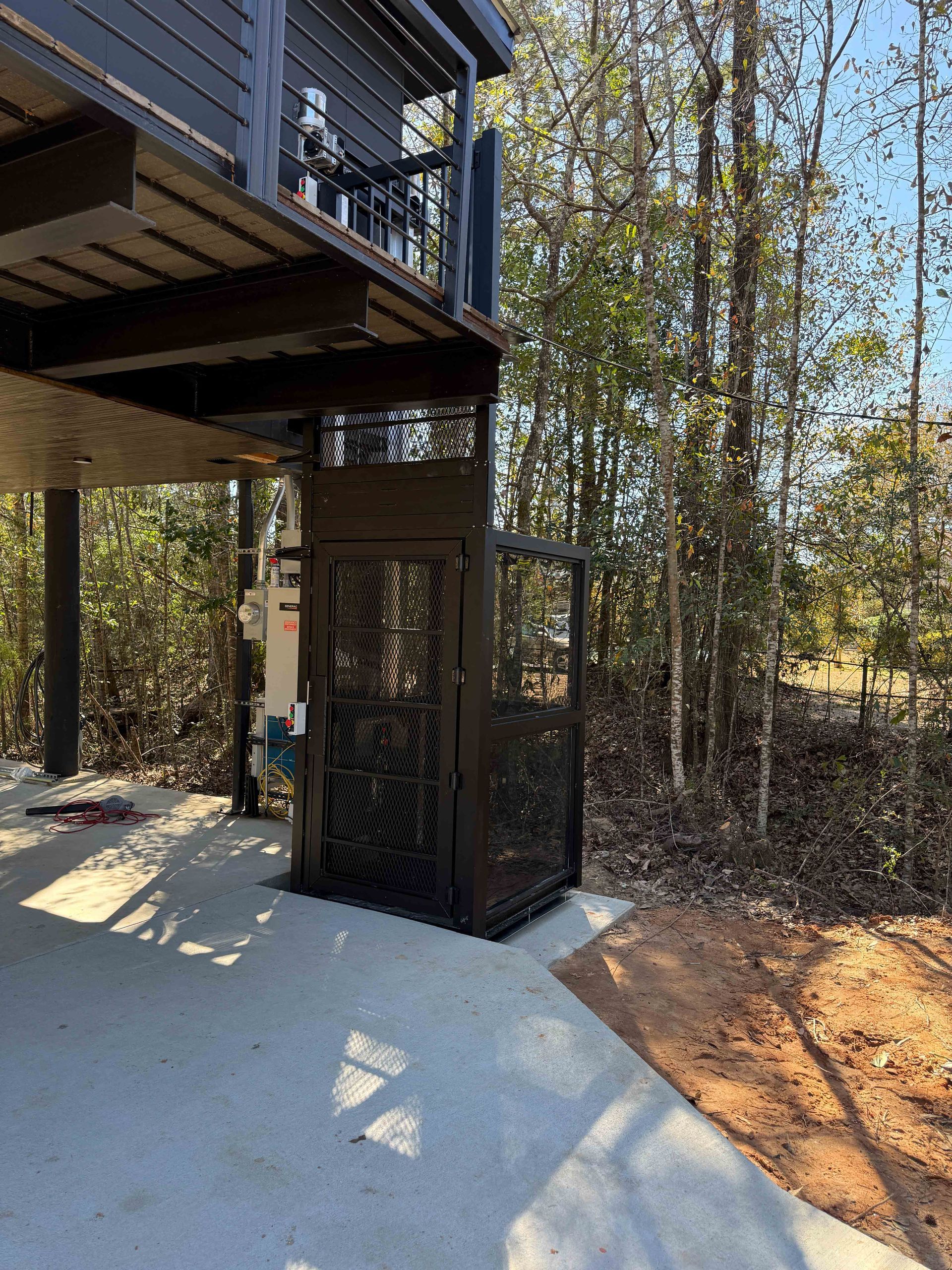 Stairway landing under a raised deck beside a wooded yard, with black railings and concrete below.