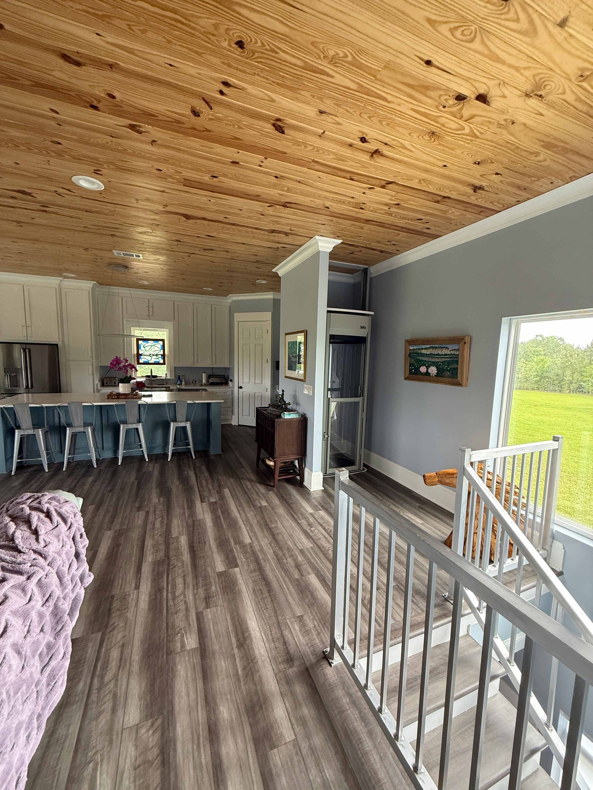 Open-plan home interior with wood ceiling, gray walls, kitchen island, and stair railing by a bright window