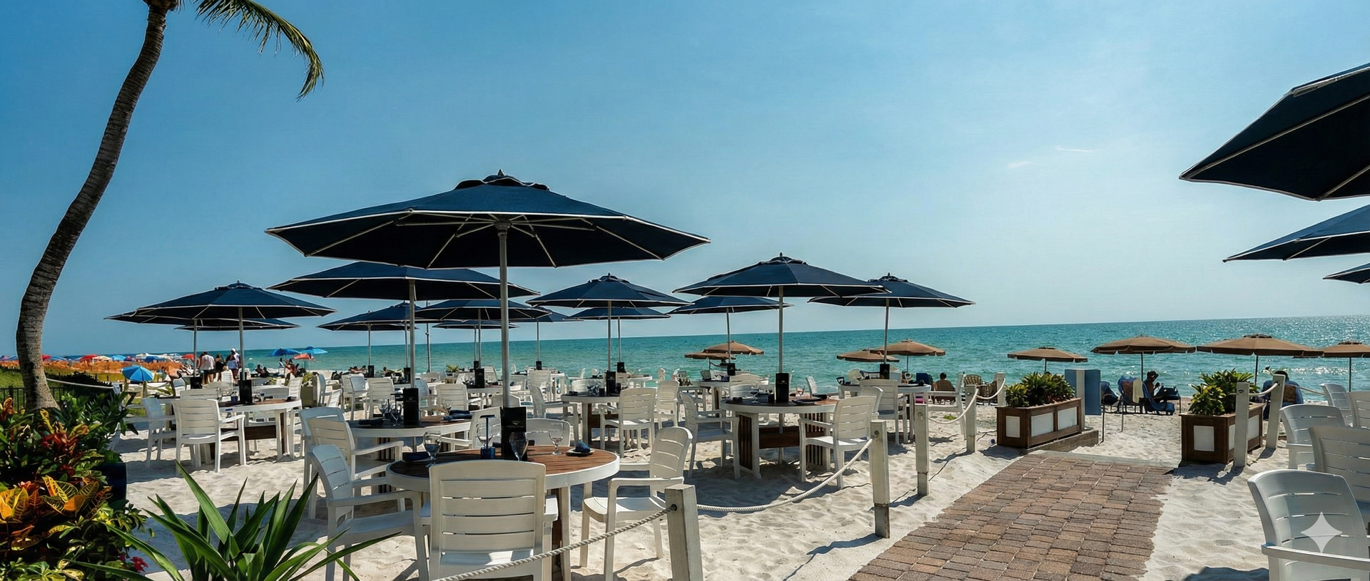 Beachside restaurant with tables and umbrellas on sand overlooking the ocean.