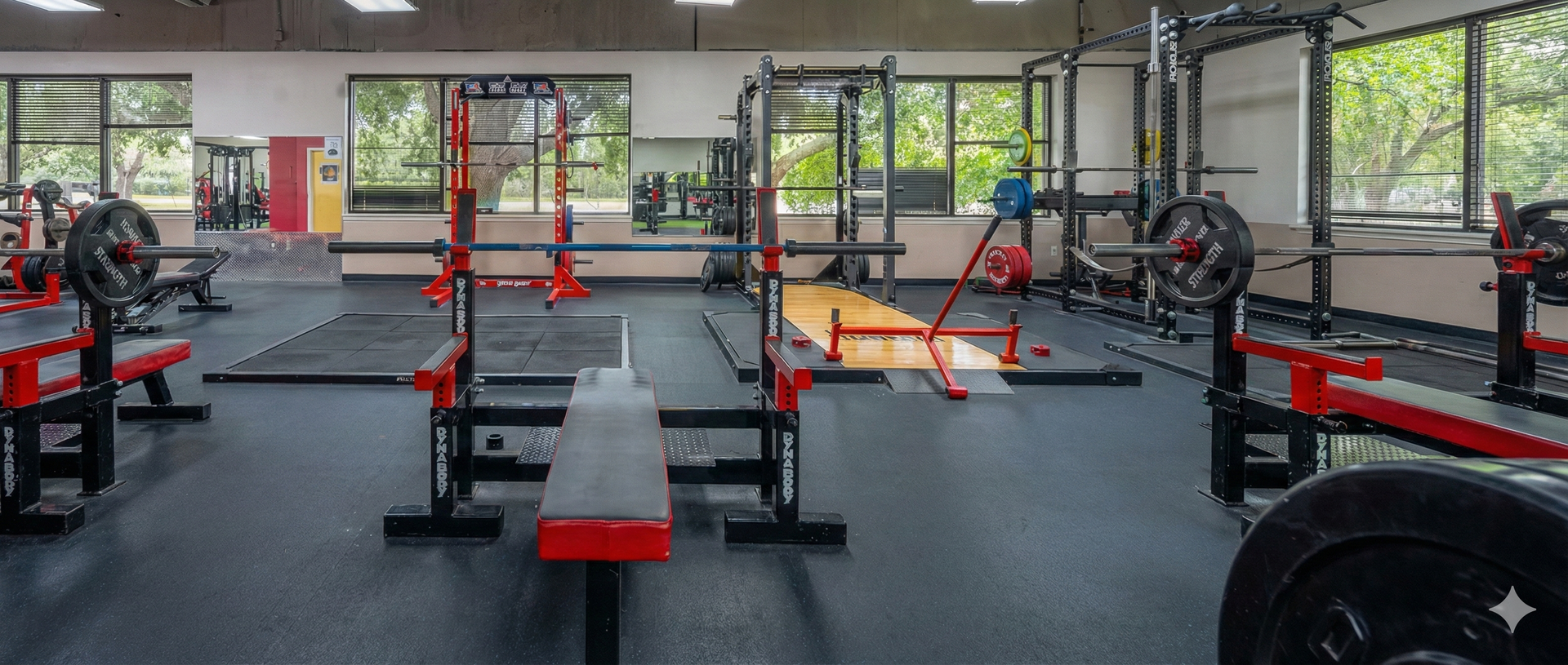 Weightlifting gym with benches, racks, and weights, windows in the background.