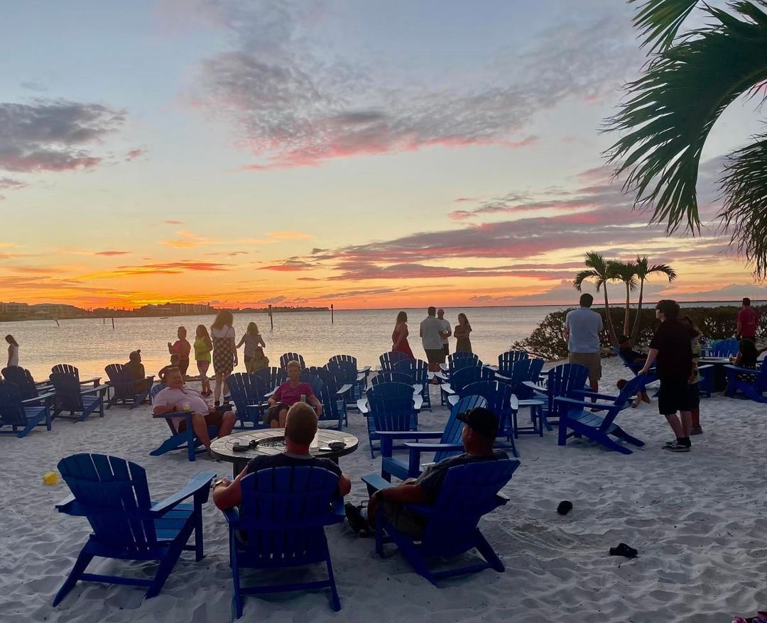 Beachgoers watch sunset over water; blue chairs on sand, palm tree, colorful sky.