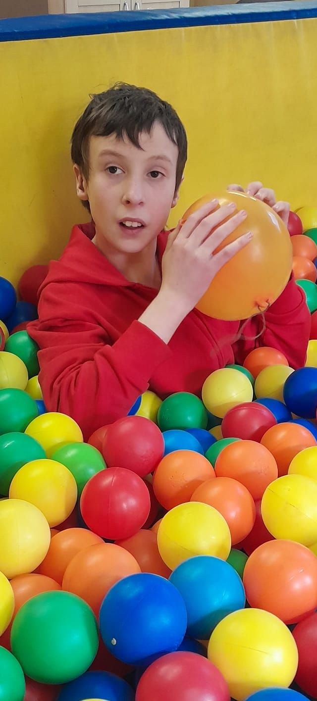 A young boy is playing in a ball pit holding an orange balloon.