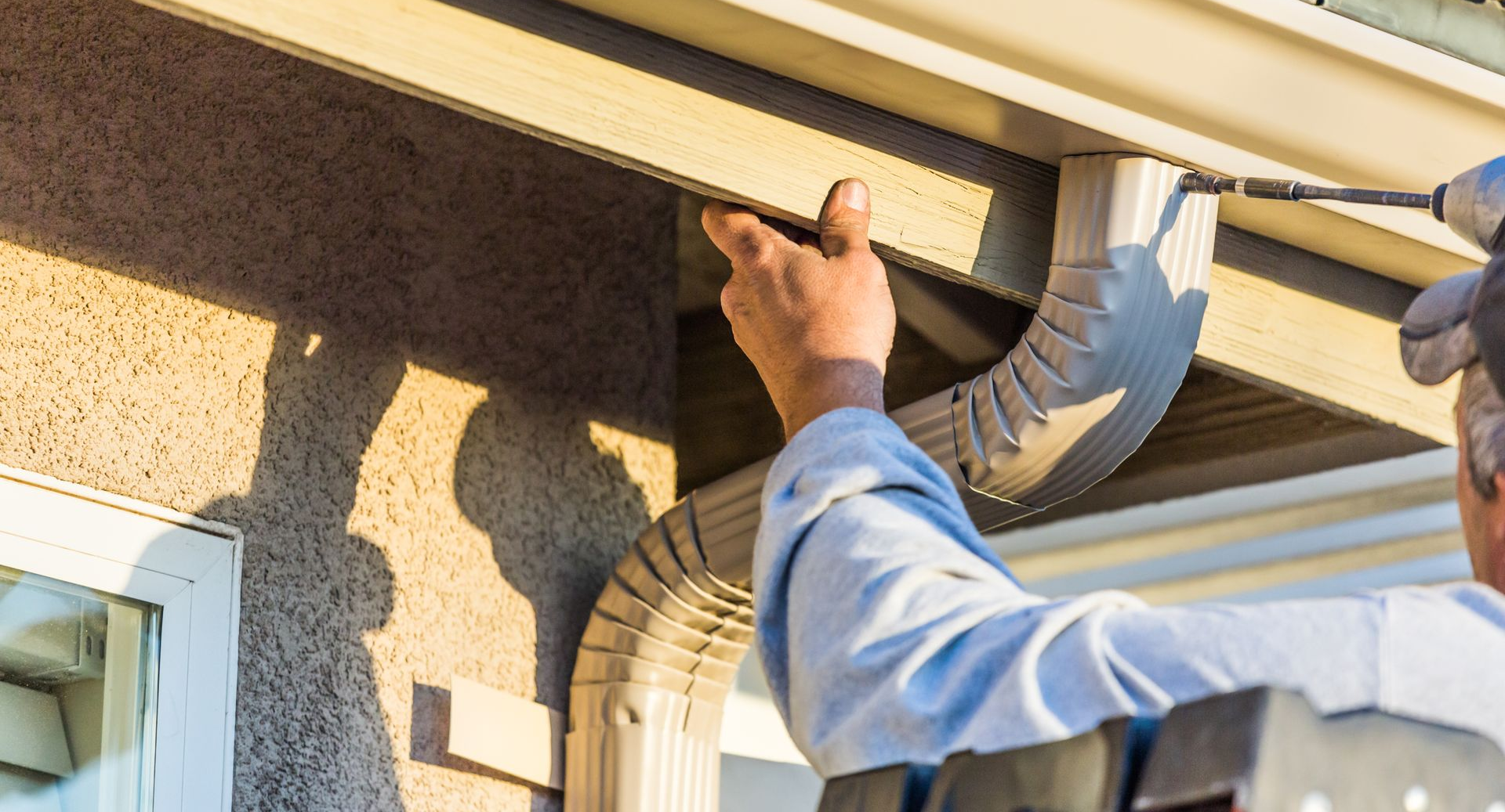 Worker installing guttering on a house roof, holding a tool near a downspout.
