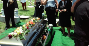 Flower-adorned black casket at an outdoor funeral, with mourners standing nearby on green turf.